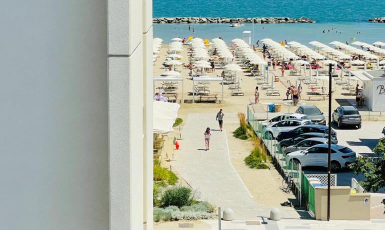 Plage bondée avec parasols blancs et mer bleue, vue entre les bâtiments.