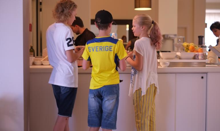 Tre bambini parlano in cucina, uno indossa una maglia gialla con scritto 'Sverige'.